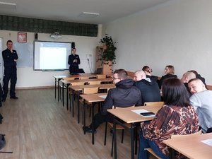 Na zdjęciu widoczna jest policjantka oraz policjant, którzy są zwróceni twarzą do młodzieży siedzącej w ławkach szkolnych i biorących udział w zajęciach.