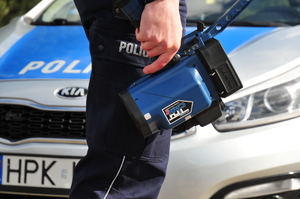 Policjant trzymający ręczny miernik prędkości w ręce wzdłuż nogi. W tle radiowóz.