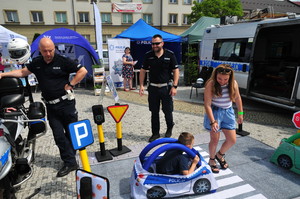 Policjant stojący przy motocyklu. Drugi policjant podczas zabawy z dziewczynką i chłopczykiem korzystającym z policyjnego auto chodzika. Obok radiowóz, a w tle policyjne stanowisko