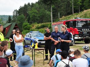 Policjantka mówiąca do dzieci. Za nią policjant, strażacy i pracownice sanepidu. W tle radiowozy i wóz strażacki.