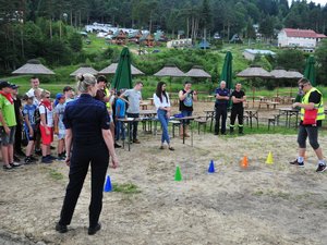 Jeden z uczestników w alkogoglach pokonuje slalom między słupkami. Obok stoi policjantka. W tle pozostali uczestnicy i organizatorzy spotkania.