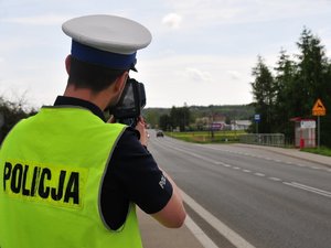 Policjant ruchu drogowego w żółtej kamizelce  stojący przy jezdni i mierzący prędkość