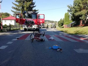 Na zdjęciu widoczne jest przejście dla pieszych, na którym leża dwa rowery, buty oraz plecak. W tle wóz strażacki