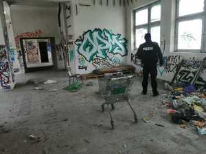 Policjant sprawdzający pustostan. Na ścianie graffiti, obok śmieci, wózek sklepowy oraz materac do spania