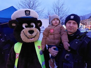 Policjant trzymający na rękach dziecko. Obok policyjny miś