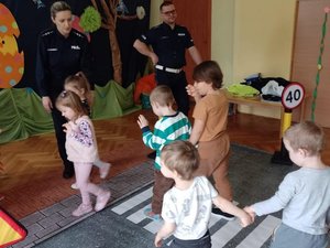 Policjanci pokazujący dzieciom jak przechodzić przez przejście dla pieszych, które jest rozłożone na podłodze