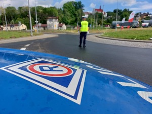 Policjant ruchu drogowego stojący w tle na rondzie. Na pierwszym planie przód radiowozu z symbolem ruchu drogowego