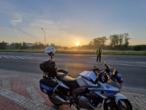 Policyjny motocykl stojący przy drodze. W tle policjant z alkomatem na środku drogi oraz wschód słońca