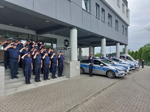 Policjanci oraz strażnicy więzienni zebrani przed komendą i oddający honor zmarłemu. Obok nich radiowozy z włączonymi sygnałami.