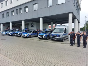 Policjanci oraz strażnicy więzienni zebrani przed komendą i oddający honor zmarłemu. Obok nich radiowozy z włączonymi sygnałami.