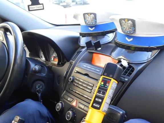 Wnętrze policyjnego radiowozu, pod szybą leżą dwie policyjne czapki i urządzenie do badania stanu trzeźwości