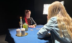 Na zdjęciu widoczne dwie siedzące przy stole frontem do siebie kobiety. Na stole widoczne zapalone świece oraz rozłożone karty.