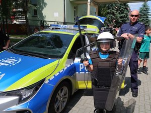 na obrazku widać chłopca ubranego w strój policyjny służący do zabezpieczeń imprez masowych, radiowóz oraz stojącego za nim policjanta