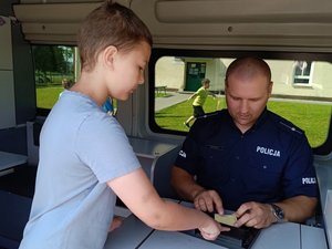 na zdjęciu widać siedzącego w radiowozie policjanta oraz chłopca odbijającego palec w tuszu do pobierania odcisków palców